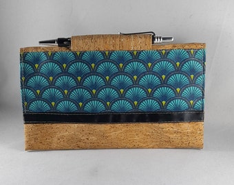 May include: A rectangular cork wallet with a teal and navy blue fan pattern fabric panel. The wallet has a black band and a pen inserted in the cork. The wallet is on a white background.