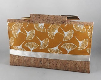 May include: A rectangular handbag with a cork base and a mustard-yellow top section. The top section features a white ginkgo leaf pattern. A silver band separates the cork base from the patterned top. The bag has a gold-colored handle.