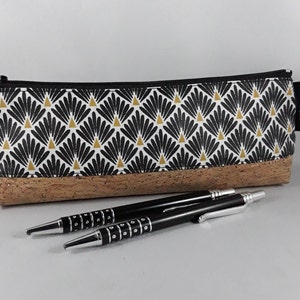 May include: A rectangular pencil case with a black zip and a tassel. The top half features a black, white, and gold fan pattern, while the bottom is made of cork. Two black pens with silver accents are in front.