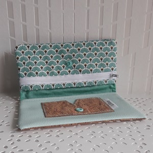 May include: Open wallet with a teal and white fan pattern on the top flap. The wallet has a white zipper, teal interior, and a cork card holder. A teal button secures the flap. The wallet is on a white surface.