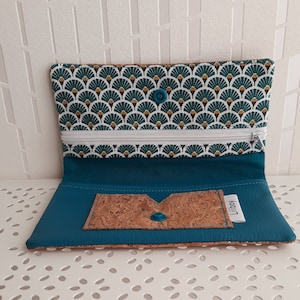 Puede incluir: Cartera teal abierta con cremallera blanca y solapa con estampado de abanico verde y blanco. El interior es teal con un detalle de corcho y cierre de botón pequeño. La cartera está abierta, mostrando su contenido.