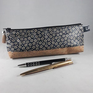 May include: A navy blue and beige floral pattern pencil case with a cork base, a black zipper, and a tassel. Two pens, one black and one gold, are next to the pencil case. The pencil case has the word "Lil" on a tag.