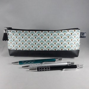 May include: A rectangular pencil case with a black bottom and a patterned top featuring a fan-like design in teal, white, and gold. Three pens in teal, silver, and black are in front of the case. The case has a black tassel and the word "Lili" on a tag.