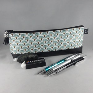 May include: A rectangular pencil case with a black base and a patterned top featuring teal and gold fan-like designs. A black tassel hangs from the zipper. Also shown are an eraser, and three pens.