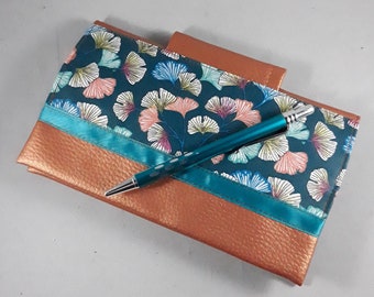 May include: A teal and copper-colored journal with a pen. The journal features a teal fabric with a floral pattern and a copper-colored leather-like material. A teal pen with white dots rests on top of the journal.