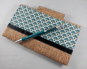 May include: A cork notebook with a teal and white fan pattern on the cover, accented by a black band. A teal pen with silver accents rests on the notebook. The notebook is made of cork material.