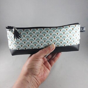 May include: A rectangular pencil case with a black zipper and tassel. The top portion features a white fabric with a teal and gold fan pattern. The bottom is black. The word "Lil" is printed on a small black tag.