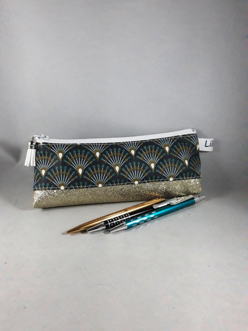 May include: A rectangular pencil case with a fan-like pattern in teal, gold, and white. The bottom section is gold glitter. A gold and a teal pen are in front of the case. The case has a white zipper and a white tassel.