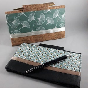 May include: Two handmade cork and fabric wallets. One is a rectangular clutch with a teal background and white leaf pattern. The other is a black wallet with a fan-shaped pattern and a pen resting on top.
