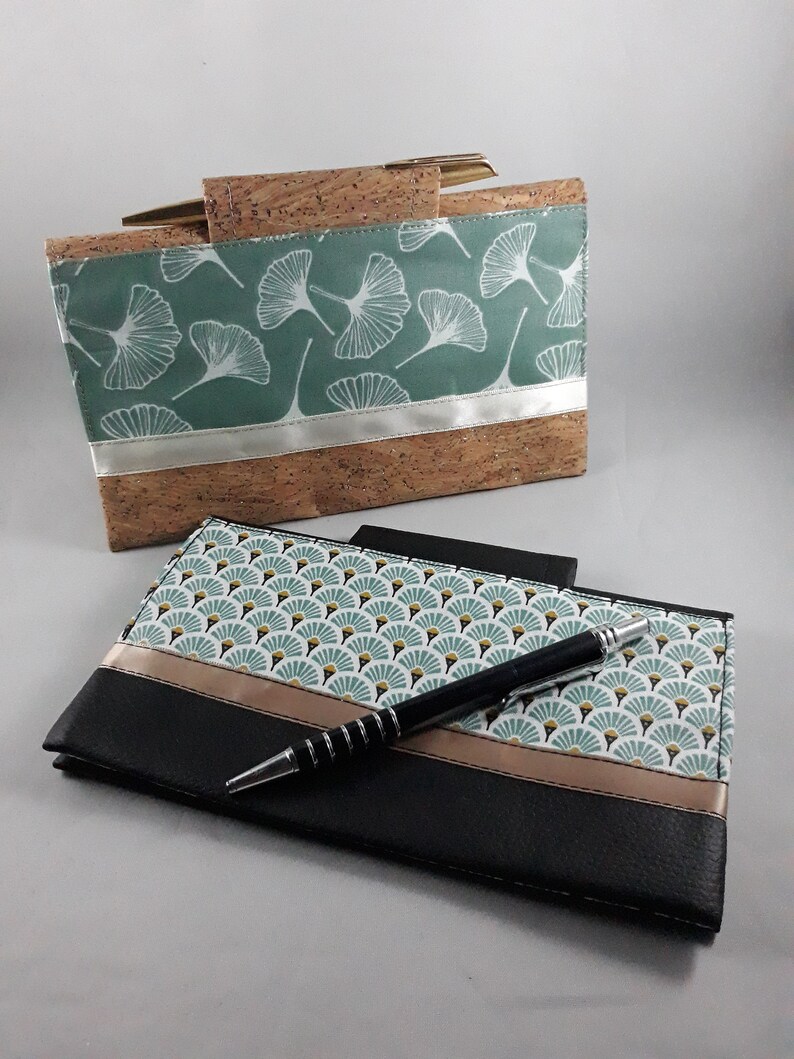 May include: Two handmade cork and fabric wallets. One is a rectangular clutch with a teal background and white leaf pattern. The other is a black wallet with a fan-shaped pattern and a pen resting on top.