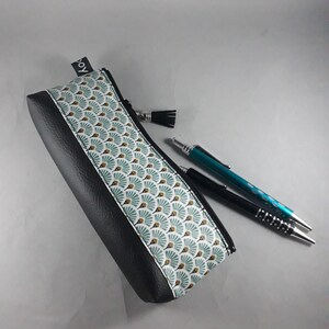 May include: A black and patterned pencil case with a zipper and a tassel. The case features a geometric fan design in teal, white, and gold. Two pens, one black and one teal, are next to the case.