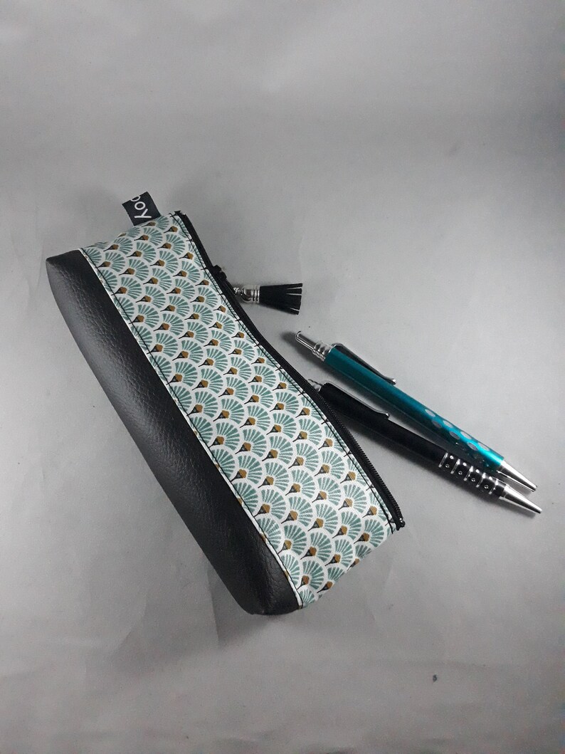 May include: A black and patterned pencil case with a zipper and a tassel. The case features a geometric fan design in teal, white, and gold. Two pens, one black and one teal, are next to the case.