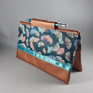 May include: A handmade checkbook cover with a teal and copper color scheme. The cover features a teal background with a ginkgo leaf pattern, copper-colored trim, and a teal ribbon accent. A pen is inserted into the top.