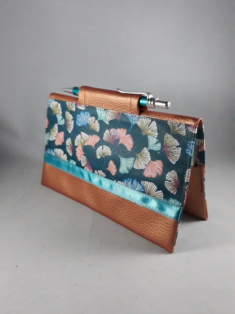 May include: A handmade checkbook cover with a teal and copper color scheme. The cover features a teal background with a ginkgo leaf pattern, copper-colored trim, and a teal ribbon accent. A pen is inserted into the top.