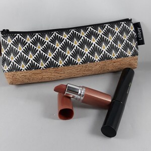 May include: A rectangular make-up bag with a black zip and tassel. The bag has a black, white, and gold geometric pattern on the top half and a cork base. A lipstick and a mascara tube are next to the bag.