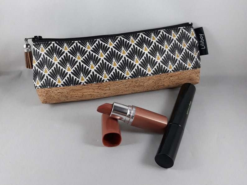 May include: A rectangular make-up bag with a black zip and tassel. The bag has a black, white, and gold geometric pattern on the top half and a cork base. A lipstick and a mascara tube are next to the bag.