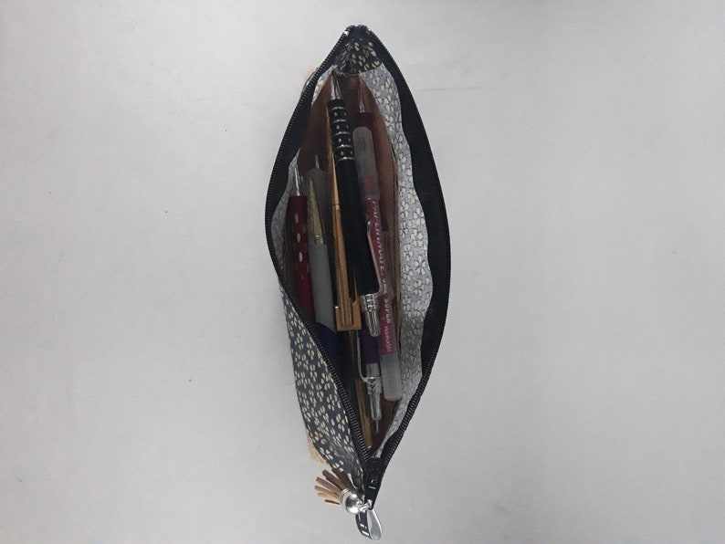 May include: A navy blue pencil case with a black zipper, filled with pens and pencils. The case has a pattern of small white and yellow dots. The interior lining is a light gray with a white floral pattern.