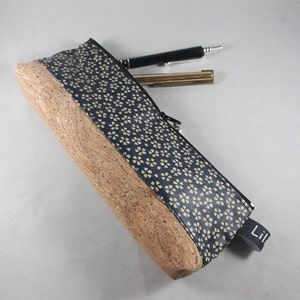 May include: A rectangular pencil case with a navy blue floral pattern and a cork base. A black pen and a gold-colored pen are next to the case. The case has a black zipper and a small black tag that says "Lil".