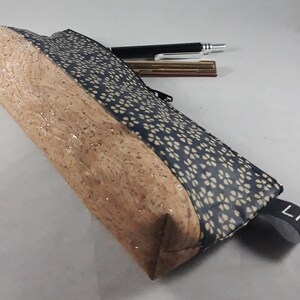 May include: A rectangular pencil case with a natural cork exterior and a navy blue fabric panel with a white floral pattern. A black zipper and a small black fabric loop with the word "Lili" are visible. A pen and a metal pen are on top.