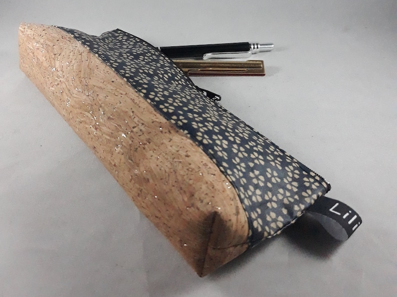 May include: A rectangular pencil case with a natural cork exterior and a navy blue fabric panel with a white floral pattern. A black zipper and a small black fabric loop with the word "Lili" are visible. A pen and a metal pen are on top.