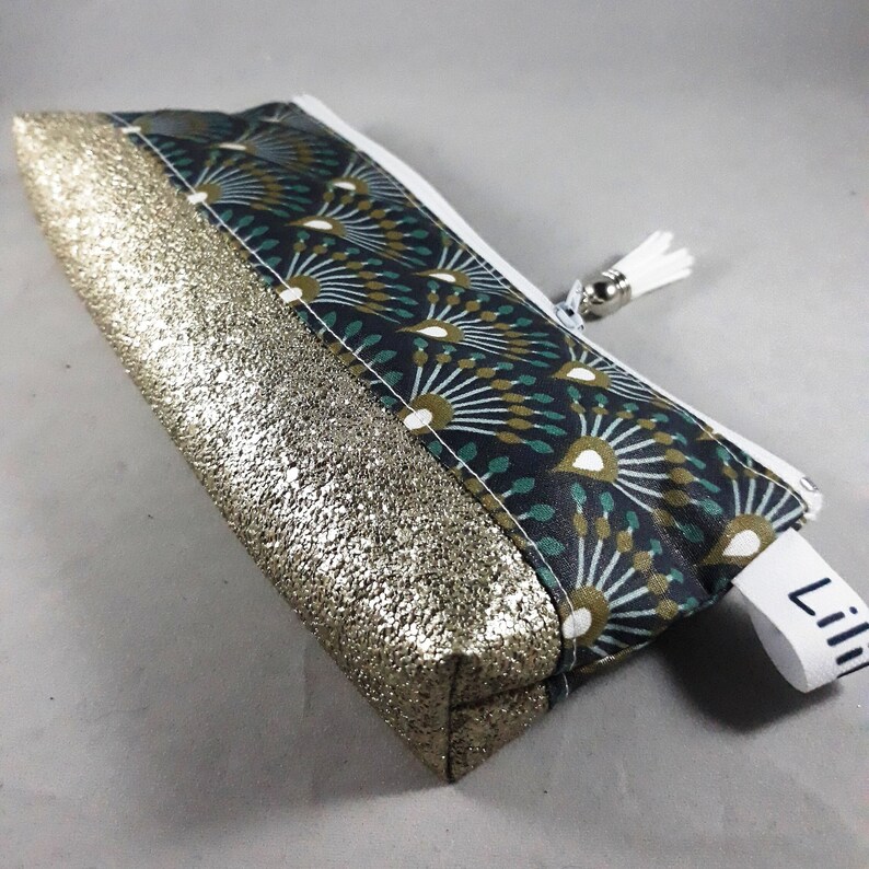 May include: A rectangular pencil case with a gold glitter fabric on the bottom half and a patterned fabric on the top half. The patterned fabric has a dark teal background with a white, teal, and gold fan design. A white zipper and tassel complete the design.