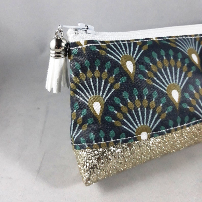 May include: A small, zippered pouch with a dark teal and gold fan pattern. The pouch has a gold metallic bottom and a white zipper. A white tassel hangs from the zipper pull.