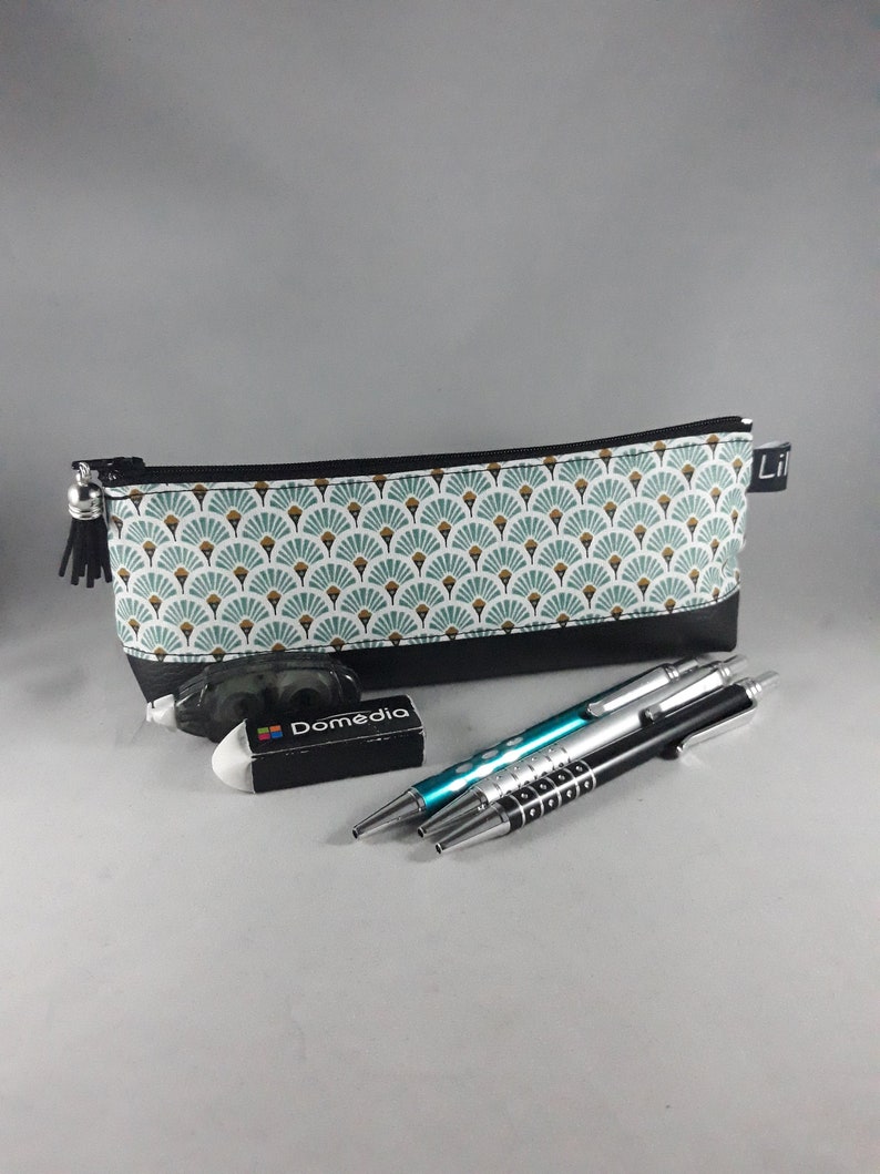 May include: A rectangular pencil case with a black base and a patterned top featuring teal and gold fan-like designs. A black tassel hangs from the zipper. Also shown are an eraser, and three pens.