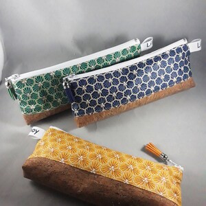 May include: Three pencil cases with geometric patterns and cork bases. One is yellow with white and gold, another is green with gold, and the third is navy blue with gold. Each has a zipper and a tassel.