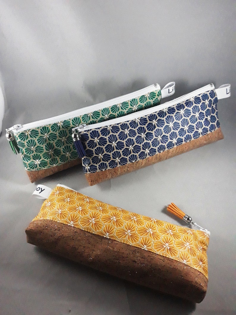 May include: Three pencil cases with geometric patterns and cork bases. One is yellow with white and gold, another is green with gold, and the third is navy blue with gold. Each has a zipper and a tassel.