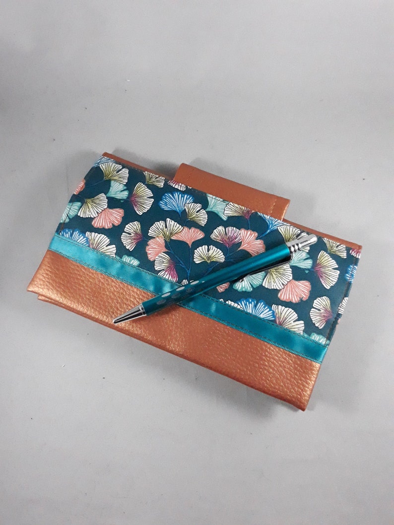 May include: A teal and copper-colored journal with a pen. The journal features a teal fabric with a floral pattern and a copper-colored leather-like material. A teal pen with white dots rests on top of the journal.