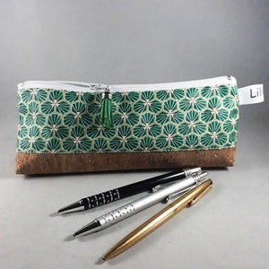 May include: A green and gold geometric pattern pencil case with a cork base, a white zipper, and a green tassel. Three pens in black, silver, and gold are in front of the case.