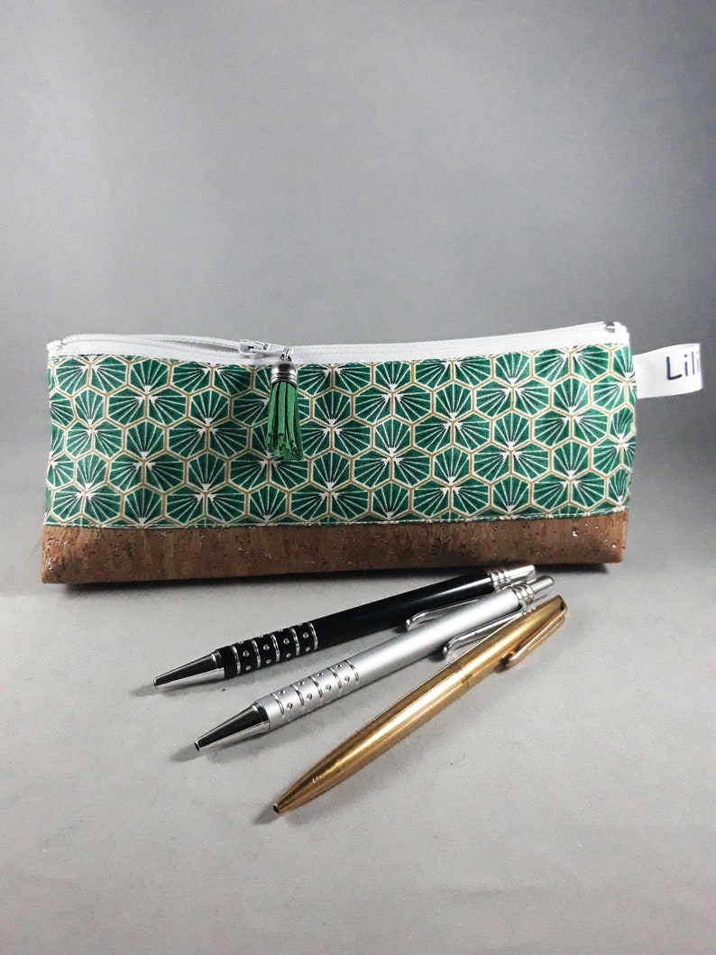 May include: A green and gold geometric pattern pencil case with a cork base, a white zipper, and a green tassel. Three pens in black, silver, and gold are in front of the case.