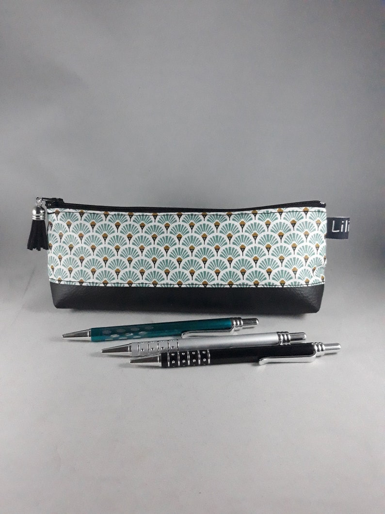 May include: A rectangular pencil case with a black bottom and a patterned top featuring a fan-like design in teal, white, and gold. Three pens in teal, silver, and black are in front of the case. The case has a black tassel and the word "Lili" on a tag.