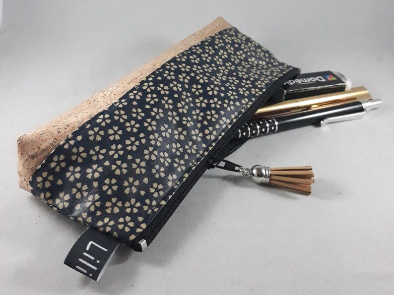 May include: A rectangular pencil case with a black zipper and a tassel. The top section is made of cork, while the main body features a navy blue floral pattern. The case is partially open, revealing pens and a pencil.