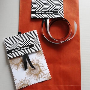 Op de afbeelding: Een oranje papieren cadeauzak met twee labels en een lint. De labels hebben een zwart-wit geometrisch patroon en de tekst "LILIBOY cr&eacute;ations". E&eacute;n label heeft een zonnebloemontwerp. Een zwart en zilver lint is op de tas gekruld.