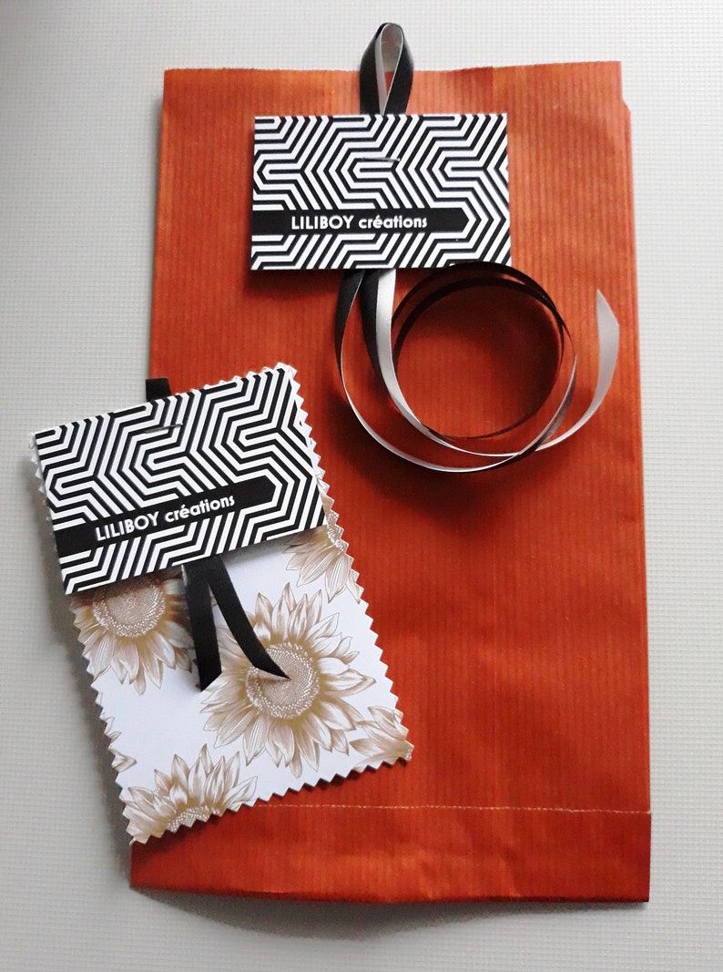Op de afbeelding: Een oranje papieren cadeauzak met twee labels en een lint. De labels hebben een zwart-wit geometrisch patroon en de tekst "LILIBOY cr&eacute;ations". E&eacute;n label heeft een zonnebloemontwerp. Een zwart en zilver lint is op de tas gekruld.