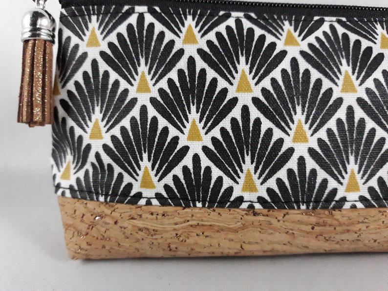 May include: A rectangular pouch with a black zip and a brown tassel. The pouch features a white fabric with a repeating black and gold fan-like pattern. The base is made of cork material.