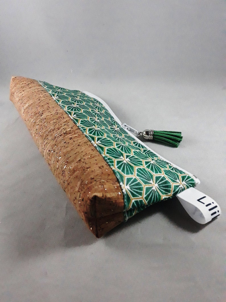 May include: A rectangular pencil case with a cork and geometric patterned design. The case features a brown cork body with a green and gold geometric pattern on the top. A white zipper and green tassel complete the design. The tag reads "Lili".