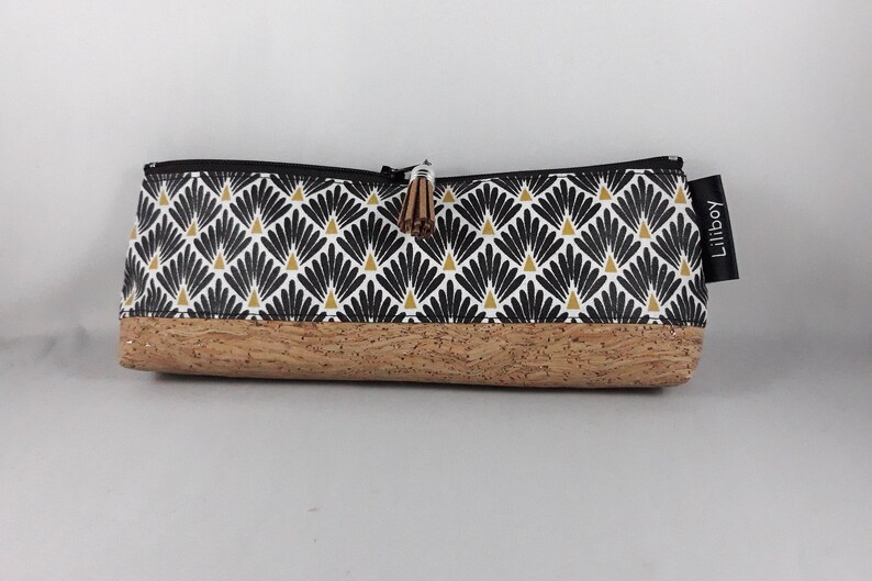 May include: A rectangular pencil case with a black zip and a cork base. The upper section has a repeating black and gold fan pattern on a white background. A small tassel and a tag reading "Liliboy" are attached.