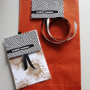 May include: An orange paper gift bag with two tags and a ribbon. The tags have a black and white geometric pattern and the text "LILIBOY cr&eacute;ations". One tag features a sunflower design. A black and silver ribbon is curled on the bag.