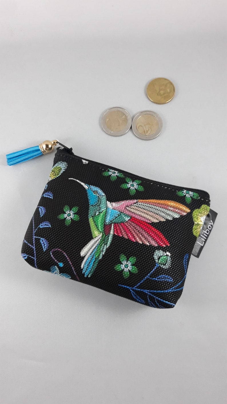 Op de afbeelding: Zwarte portemonnee met een kleurrijk geborduurde kolibrie. De kolibrie heeft blauwe, groene en rode details. De portemonnee heeft een blauwe kwast en het merklabel "Liliboy".