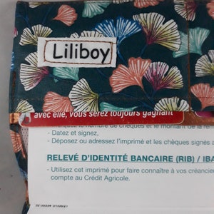 May include: A fabric wallet with a dark teal background and a pattern of stylized ginkgo leaves in shades of pink, yellow, and blue. The name "Liliboy" is stitched on a white patch.