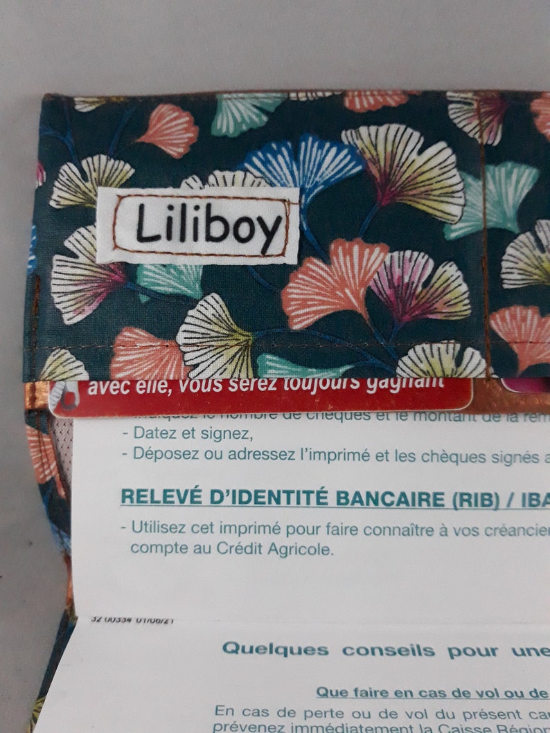 May include: A fabric wallet with a dark teal background and a pattern of stylized ginkgo leaves in shades of pink, yellow, and blue. The name "Liliboy" is stitched on a white patch.