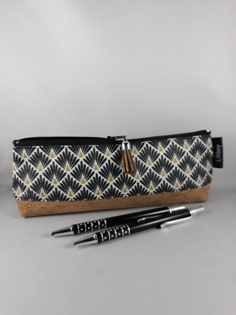 May include: A patterned pencil case with a black and white design and a gold tassel zipper pull. The case has a cork base and is paired with two black pens with silver accents. The case is approximately 20 cm long.