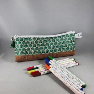 May include: A green and white patterned pencil case with a cork base and a white zipper. A green tassel hangs from the zipper pull. Several colorful markers are next to the pencil case. The pencil case has the name "Lili" on a white tag.