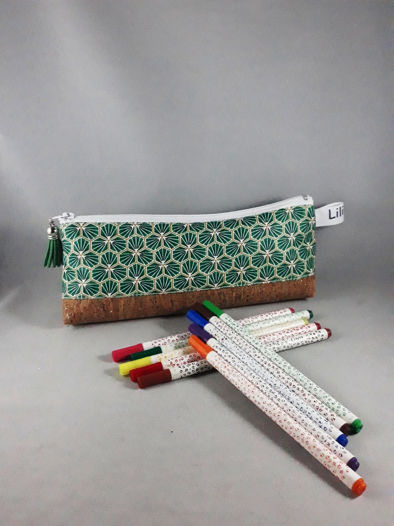 May include: A green and white patterned pencil case with a cork base and a white zipper. A green tassel hangs from the zipper pull. Several colorful markers are next to the pencil case. The pencil case has the name "Lili" on a white tag.