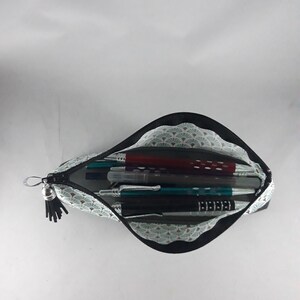 May include: Open pencil case with a black zipper and tassel. The interior features a light blue and white fan pattern, holding various pens and pencils. The case is open, displaying the contents against a white background.