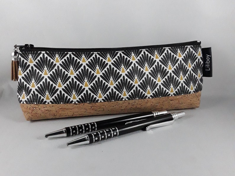 May include: A rectangular pencil case with a black zip and a tassel. The top half features a black, white, and gold fan pattern, while the bottom is made of cork. Two black pens with silver accents are in front.