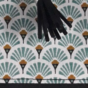May include: Close-up of a patterned fabric with a black tassel. The fabric features a repeating design of teal fan shapes with gold accents on a white background. A black zipper and a silver metal zipper pull are visible.