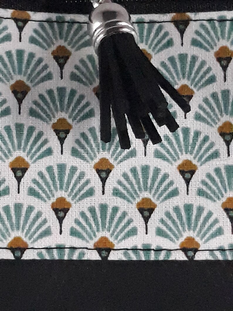 May include: Close-up of a patterned fabric with a black tassel. The fabric features a repeating design of teal fan shapes with gold accents on a white background. A black zipper and a silver metal zipper pull are visible.